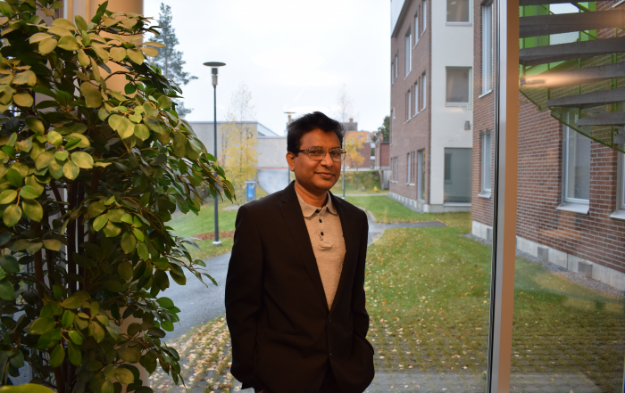 SM Shafiqul Alam seisoo ikkunan edessä ja hänen vieressään vasemmalla on suuri huonekasvi.